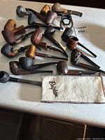 Overview of 18 briar pipes arranged on countertop with cloth sack and metal pipe holder
