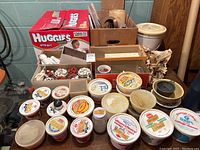 Overview of multiple containers of pottery glaze colours, stored in repurposed plastic containers and cardboard boxes including a Huggies box.