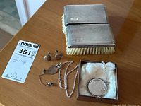 Full lot photo showing two rectangular sterling silver brushes with bristles, chain necklace with coin pendant, three screw back earrings, and a tie pin arranged on a wood surface.