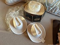 Three off-white felt cowboy hats arranged on bedding with one in original box and plastic covers