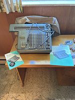 Friden automatic calculator on wooden table with beige plastic dust cover, two manuals beside it.