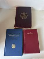 Photo of three antique books: Webster's Collegiate Dictionary (large red hardcover, worn), Record of American Uniform and Historical Buttons (blue hardcover), and Bedside Diagnostic Examination (red hardcover)