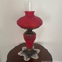 Full view photo showing the entire lamp with red satin shade, red satin body, and clear chimney on an ornate iron base.