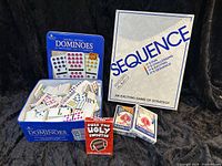 Overview of the set including Sequence board game box, open blue tin box with domino pieces, two decks of Bicycle playing cards and Pass the Ugly Sweater card game.