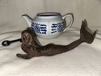 Full view of cast iron mermaid figurine lying next to small ceramic white and blue teapot and metal key on a white fabric background.