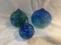 Three spherical handblown glass ornaments in green and blue tones on beige cloth background