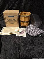 Photo of the miniature Longaberger waste basket with original box, certificate of authenticity, cloth liner in blue check pattern, plastic liner, and literature.