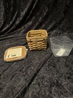 Photo showing the small woven basket, wooden lid with sticker, and clear plastic liner on a black fabric background.