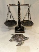 Front view of the decorative metal balance scale and wooden turtle figurine side by side on a neutral background.