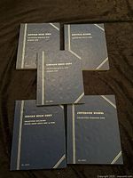 Five blue coin collection books on dark surface, showing their covers with titles and collection ranges.