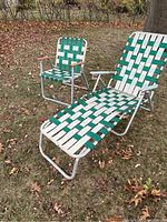 Two vintage aluminum outdoor chairs on grass and fallen leaves; lounge chair with extended leg rest and lawn chair with wooden armrests, both featuring green and white woven webbing seats.