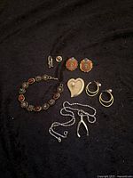 All items laid out on black fabric background: bracelet, two pairs of earrings, heart-shaped piece, chain with pendant, and pins.