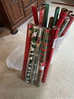Photo of 11 rolls of Christmas wrapping paper standing inside large clear plastic storage container on carpeted floor.