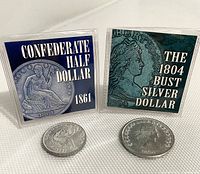 Two tribute coins displayed upright in clear plastic sleeves, showing front designs and COA cards: Confederate Half Dollar 1861 and 1804 Bust Silver Dollar copy.