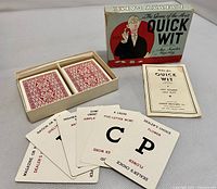 Photo of two decks of Quick Wit playing cards in plastic trays, some cards fanned out showing letter prompts and clues, with the instruction booklet nearby