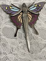Front view of silver-tone fairy pin with purple, blue, tan, and yellow enamel detailing on wings, displayed on lace background.