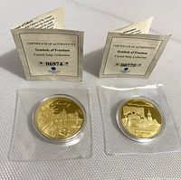 Two 24K layered gold-tone collectible coins with Swarovski crystals placed in protective cases along with their Certificates of Authenticity, showing the front design details and certificate numbers.