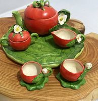 Miniature apple-themed doll tea set displayed on wood surface. Includes teapot, sugar bowl, creamer, two cups, and a leaf tray.