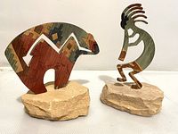 Pair of colorful flat metal figures mounted on separate stone bases. Left is bear figure with geometric southwestern pattern, right is Kokopelli figure playing flute.