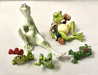Five varied frog figurines arranged on flat surface, one tall light green, one with beer can, smaller ones with hearts and other poses.