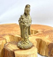 Front view of small metal Kwan Yin statuette standing on natural wood base showing figure holding child with robe details.