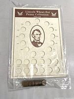Front view of Lincoln Wheat-Ear Penny Collection paper holder with designated slots for 24 coins dated 1938-1954, and visible loose pennies in a clear plastic bag below the holder.