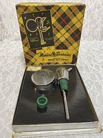 Music box, pouring spout, and rubber stopper inside original tartan box on lace background.