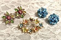 Photo of all three jewelry pieces: one floral brooch and two pairs of clip earrings (floral rhinestone and blue bead clusters) on lace background.