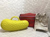 Three bags grouped together: yellow pickle hard plastic bag, red makeup bag, cream snack bag with black pattern.