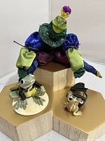 Trio of frogs including large fabric-bodied frog and two ceramic frogs, one playing piano, shown on hexagonal platforms.