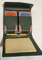Green leatherette playing card holder containing two decks of Bicentennial-themed cards and two pencils with worn paint, plus a small paper notepad, showing age and wear.