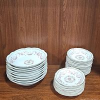 Stack of 9 soup bowls, 19 dessert bowls, all white china with pink rose design and scalloped edges, arranged on wooden surface.