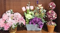 Three different floral arrangements displayed together: pink flower basket, metal container with mixed flowers, two spherical topiary flower arrangements.
