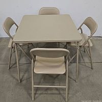 Square folding table with 4 beige folding chairs arranged around it, on concrete floor, plain white backdrop