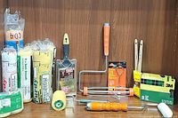 Photo showing multiple paint rollers including 12-inch and foam rollers, a wooden-handled brush, sanding blocks, and packs of roller covers on a wooden shelf.