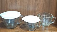 View of three large glass measuring cups, two with white lids, placed on wooden surface against wood wall background.