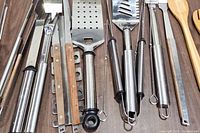 Close-up photo showing multiple metal grill utensils including spatulas, tongs, forks and skewers laid on a wood surface.