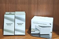 Two boxes of Sharper Image printer ink cartridges next to the Sharper Image smartphone photo printer original box.