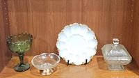 Four vintage glass serving pieces displayed on wooden shelf including milk glass deviled egg plate with gold trim, green pedestal candy dish, clear footed candy dish, and clear glass bowl with metal base and lid.