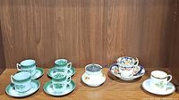 Four green floral espresso cups with matching saucers, two tea cups and saucers with the same green floral design, and one vintage floral sugar bowl with metal lid and spoon all displayed against a wooden background.