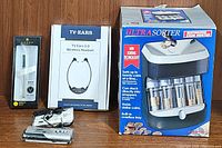 Three electronic items on wooden surface: two boxed wireless headset units, boxed coin sorter and an opened box showing contents.