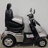 Left side profile showing metallic silver body, black padded seat with headrest, front wheel and foot platform