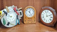 Three wooden wall clocks on shelf: building supplies saw blade clock, Mom's Kitchen clock in center, classic wood clock with Roman numerals