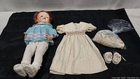 Photo showing the Walky Talky doll lying on a black cloth alongside its original clothing including a cream dress, knitted blue sweater, white shoes and socks, and a plastic bag containing additional clothing.