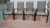 Front view of four brown metal frame patio chairs with light brown sling seats and backs arranged in a row on a wooden floor.