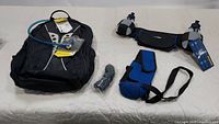 CamelBak hydration backpack with blue drinking tube, hydration belt with two bottles, a blue support strap, and a pack of squeeze tops arranged on a white surface.