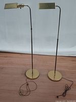 Pair of two brass Mid-Century Modern floor lamps side by side showing full height from base to lamp head, cords visible on wooden floor.