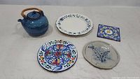 Wide shot of entire lot: blue glazed teapot with bamboo handle, dinner plate with blue floral pattern, colorful hand-painted Spanish plate, stoneware hanging plate embossed with wildflower design, and small hand-painted terra cotta tile