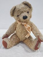 Front view of vintage Chad Valley jointed teddy bear sitting, showing plush tan fur, brown felt paw pads, and peach silk ribbon tied around neck.