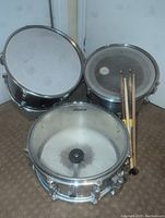 Photo showing a set of one snare drum, two tom drums, and four drumsticks placed on carpeted floor, viewed from front angle.